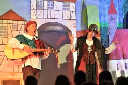 Zwei Schauspieler in mittelalterlichem Gewand singen vor gemalter Burg-Kulisse.